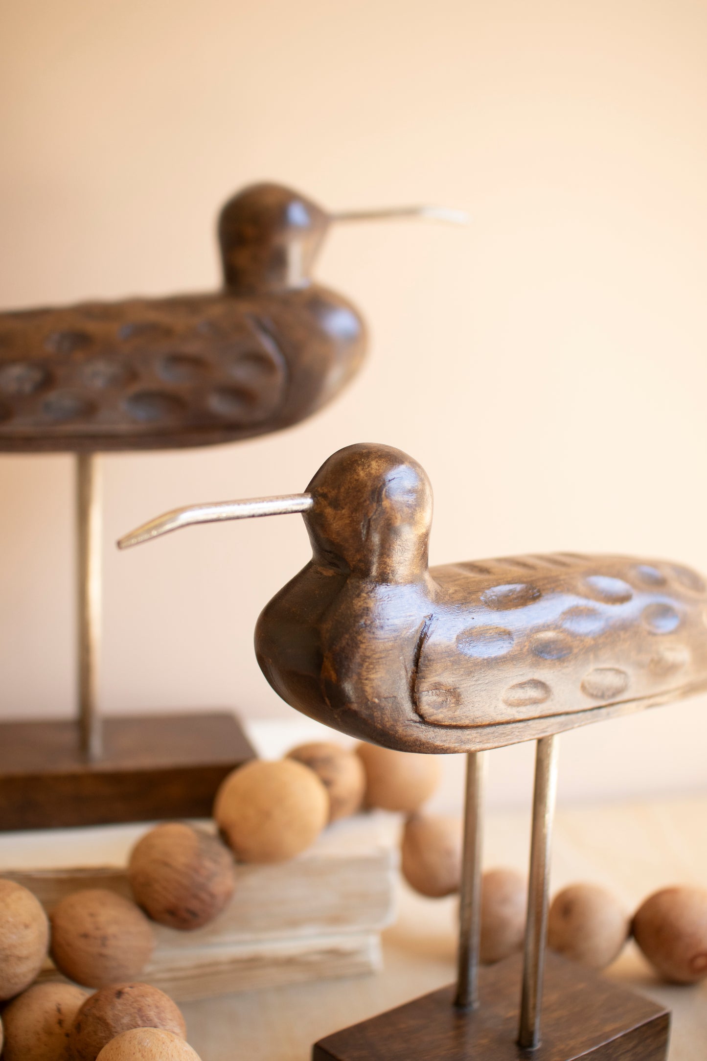 CarvedWoodenShorebirdsonStands-Setof2-9 - Decorative bronze bird sculpture on a stand with wooden beads in the background