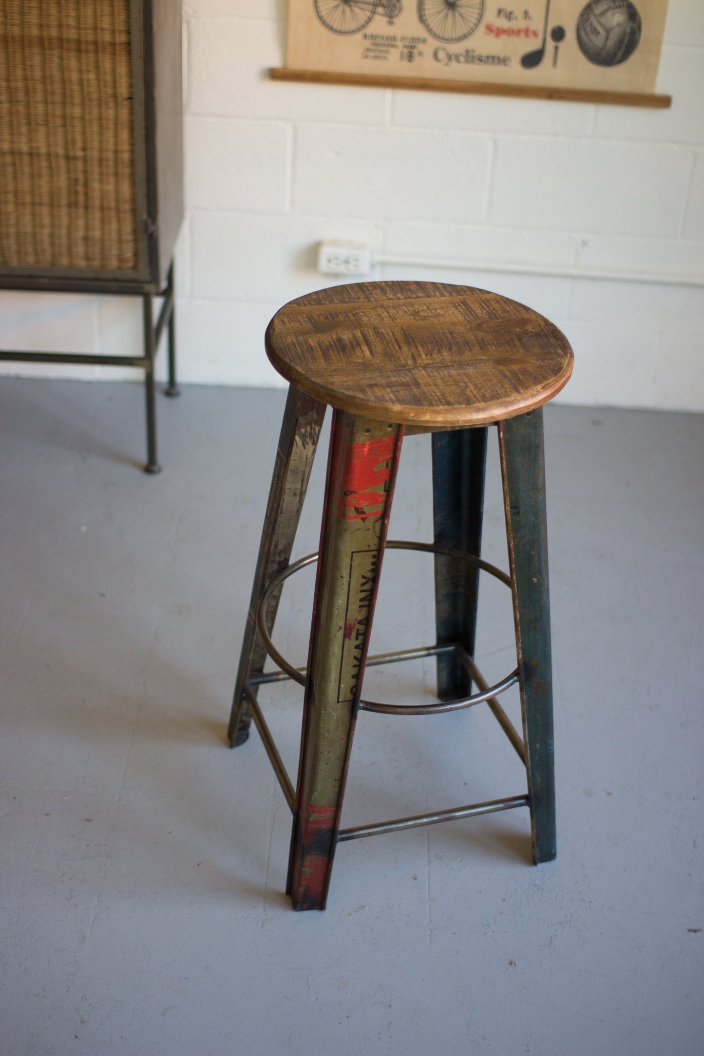 RecycledMetalBarStoolwithWoodenSeat Top View on a gray floor, with a white wall and poster in the background.
