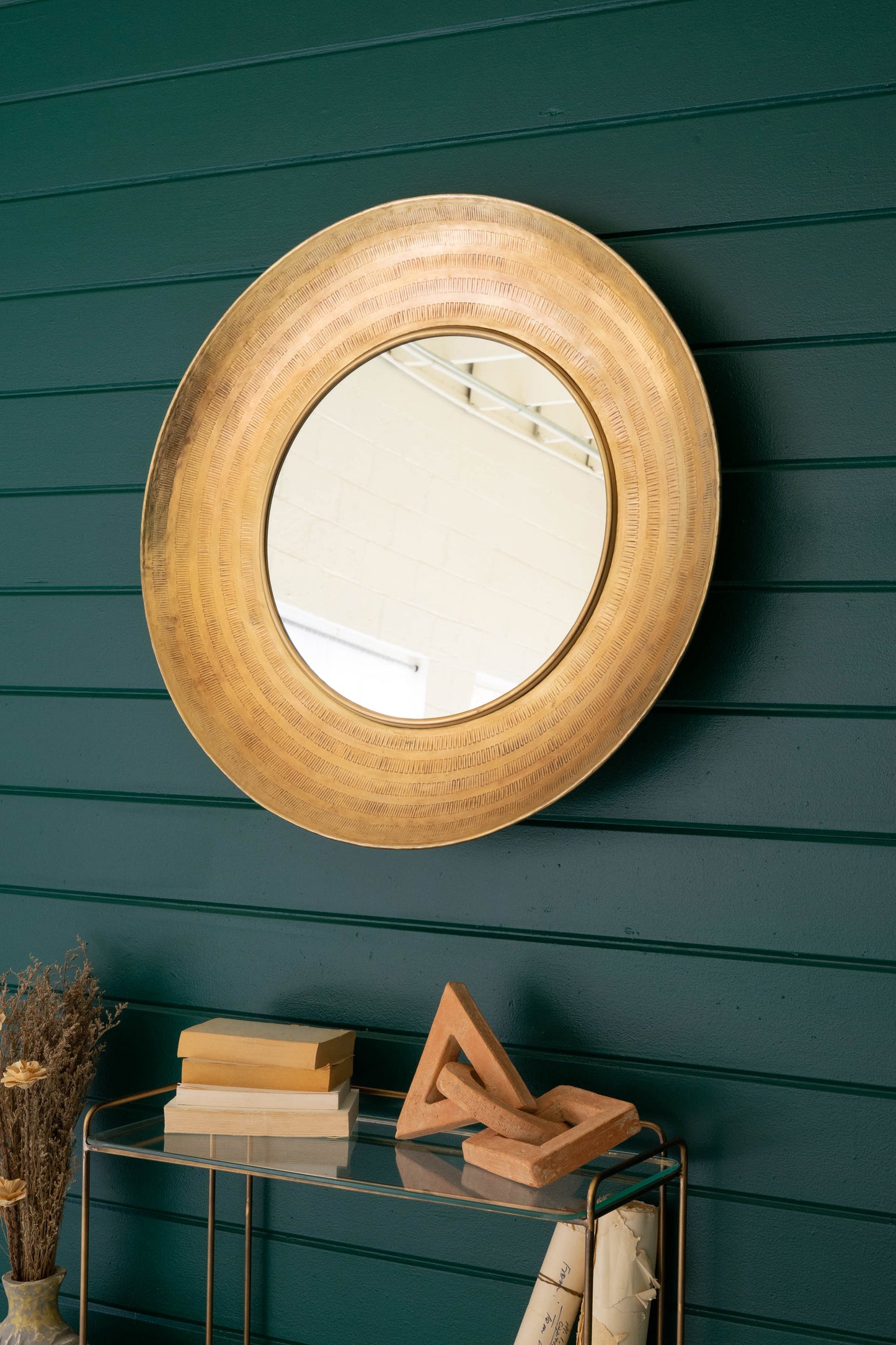 Round Hand-Hammered Antique Brass Framed Mirror reflects a room on a teal wall, framed in a warm metallic finish.