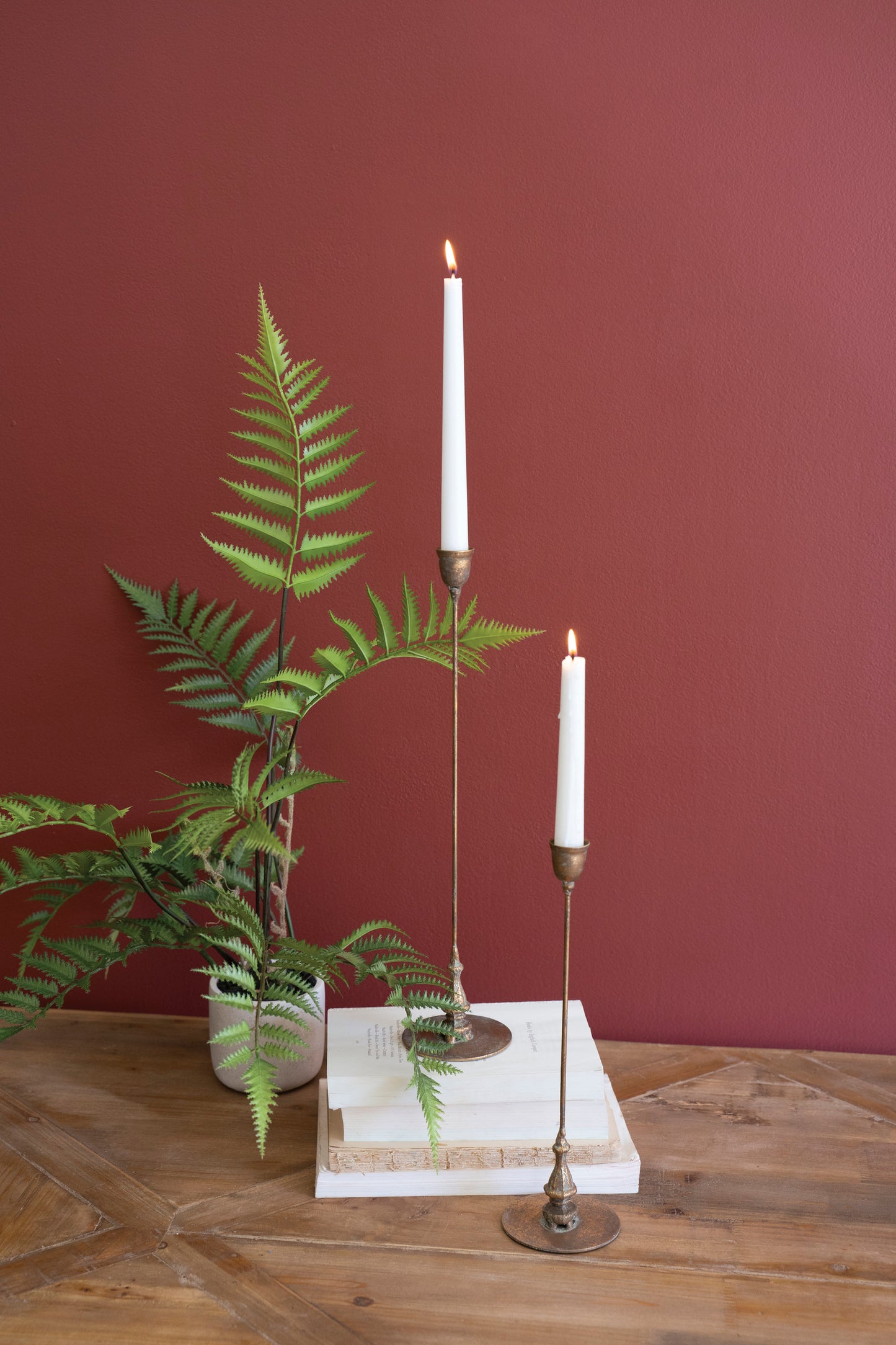antique brass candle holders by Fern plant, and books on a wooden surface with a maroon wall background
