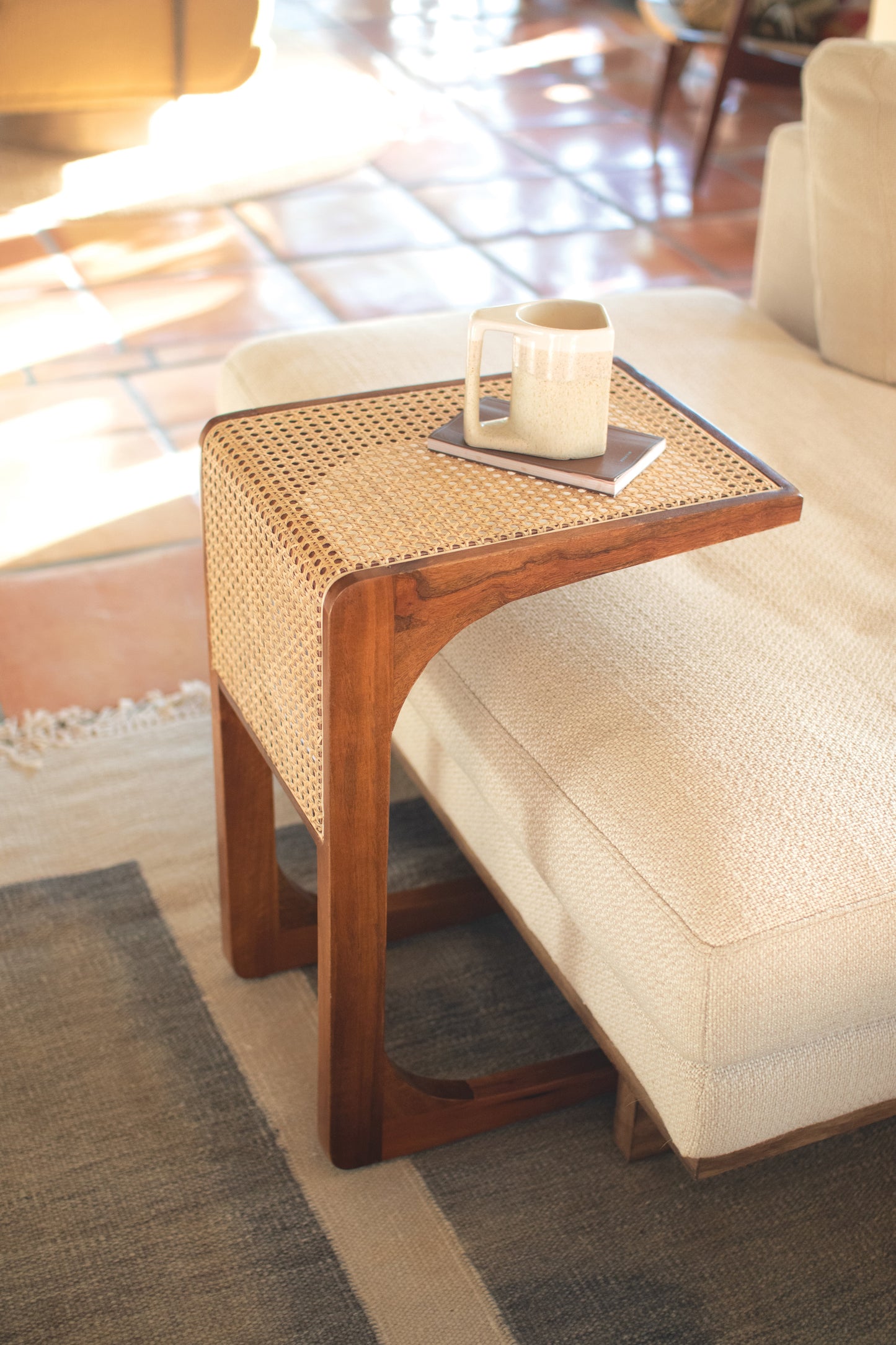 Wood Sofa Table with Woven Cane Detail beside a light-colored sofa, topped with a mug and book.