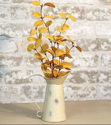 Vintage-style white watering can with yellow leaves against a textured white wall.
