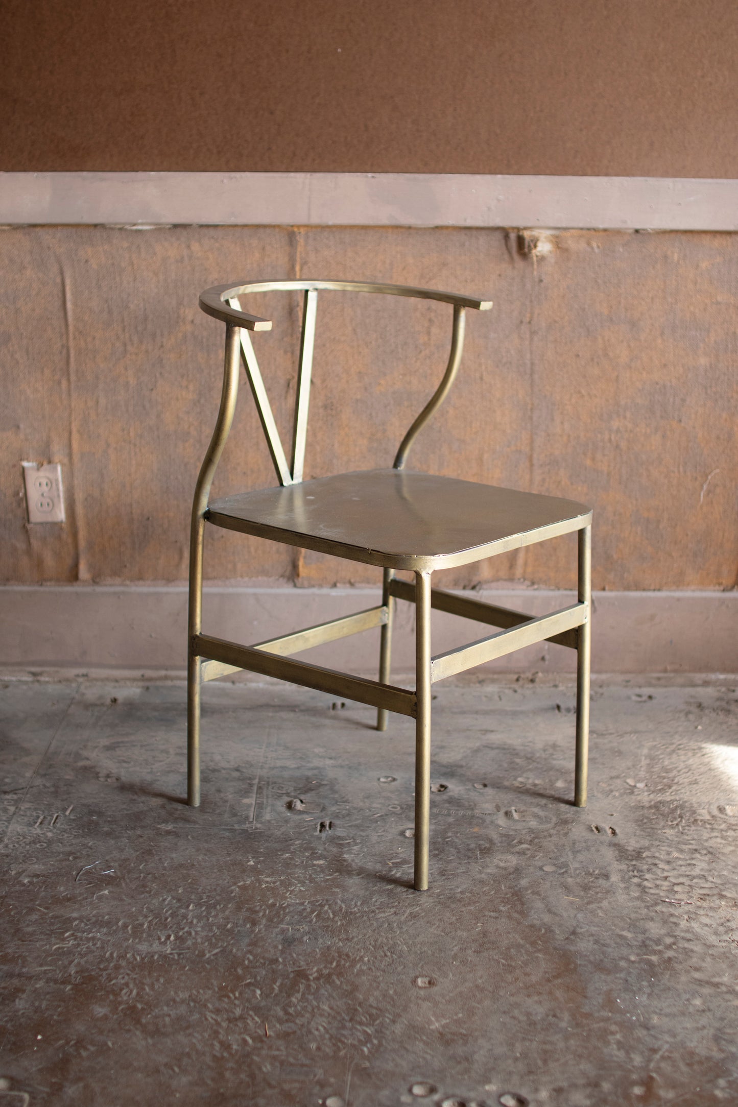 AntiqueBrassFinishMetalWishboneChair - Gold metal chair against a plain wall with concrete floor