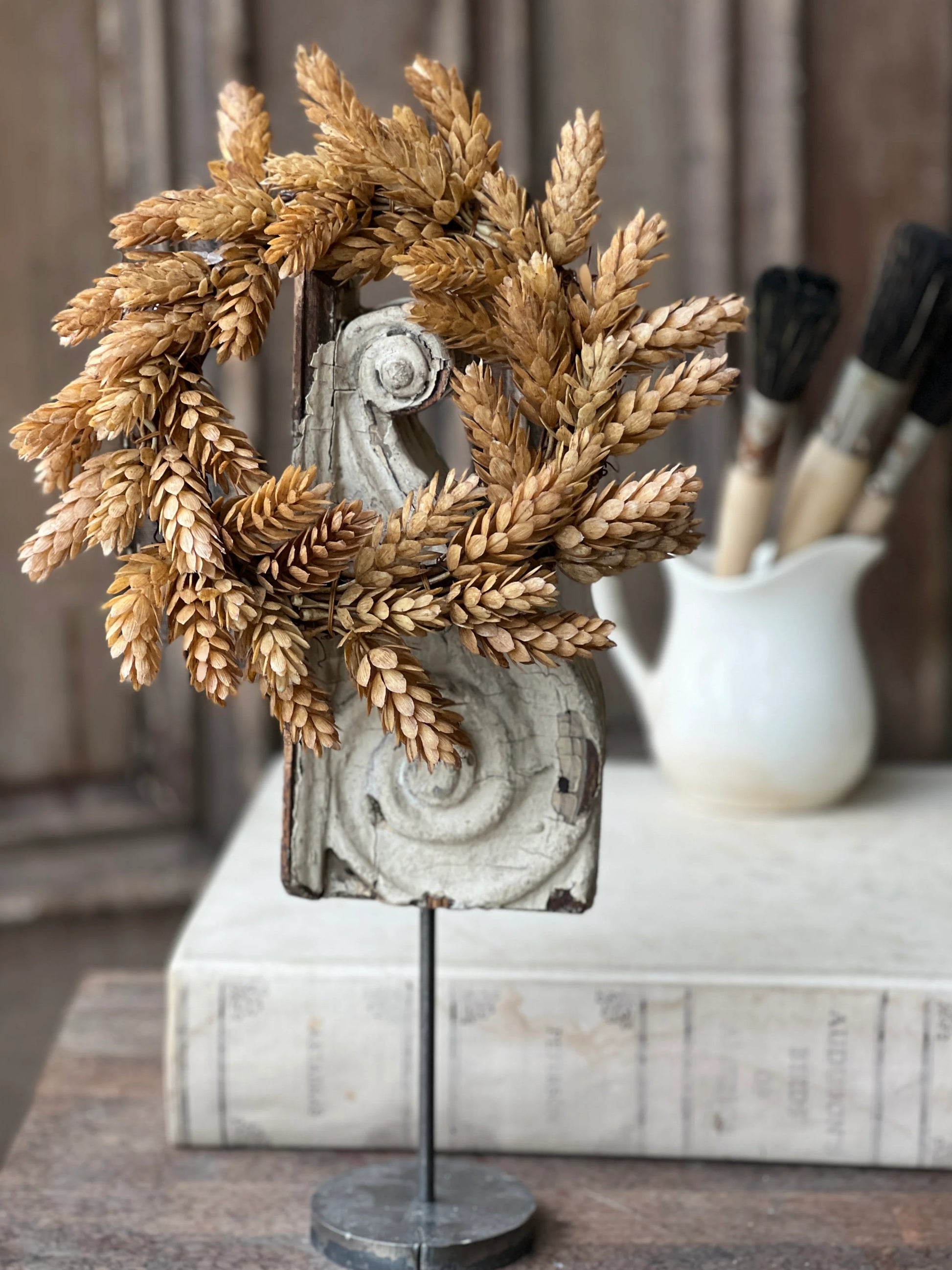 A beige hops candle ring with fall floral design, displayed on a table with a background of a workshop.