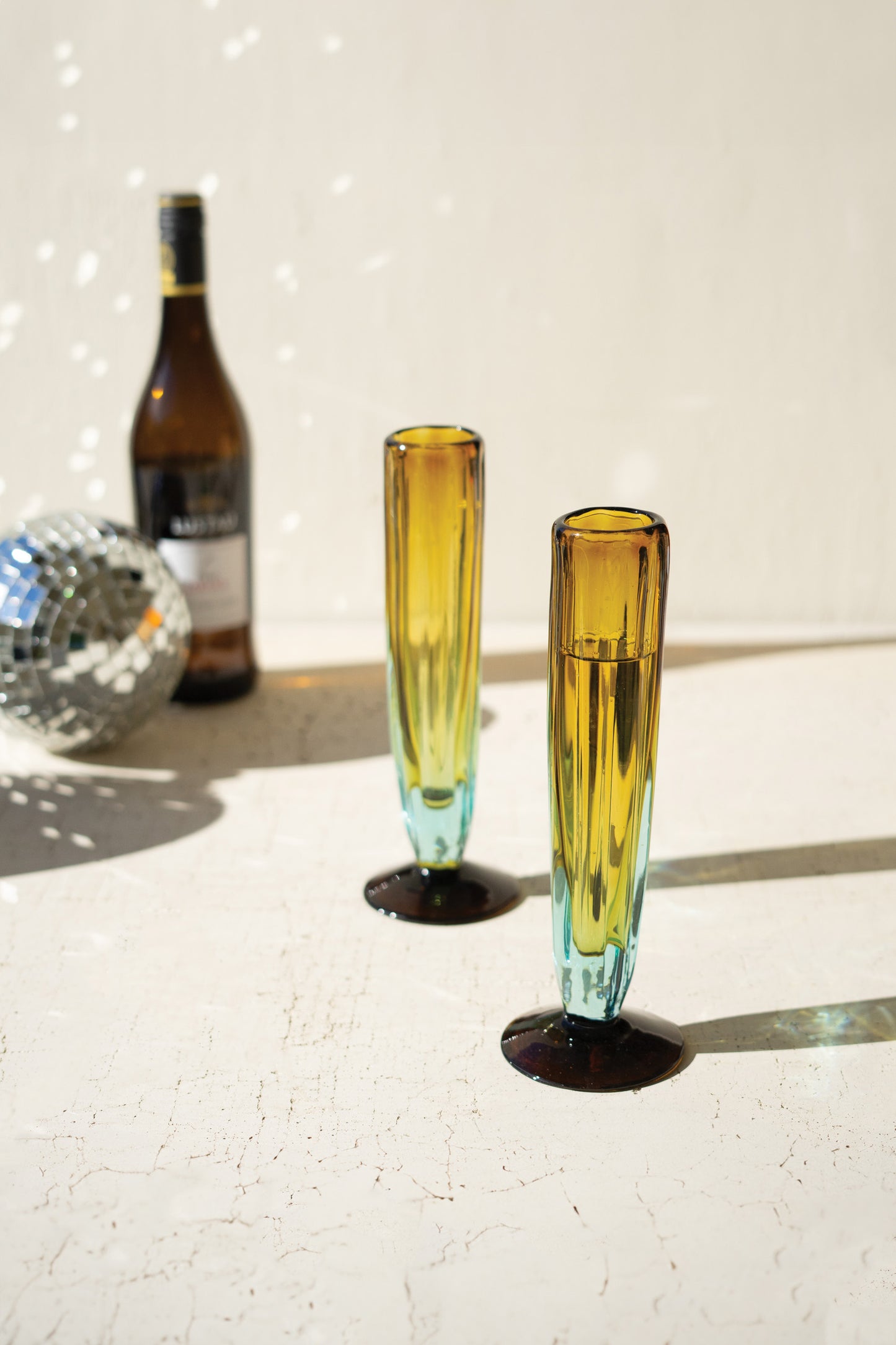 BlueandAmberRecycledChampagneGlass - Two yellow-tinted glasses on a reflective surface with a bottle and disco ball in the background.
