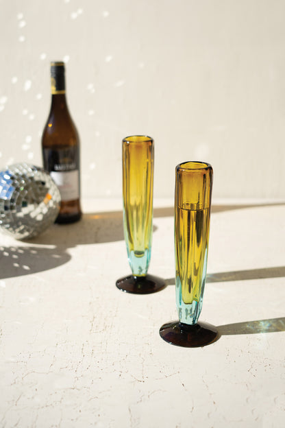 BlueandAmberRecycledChampagneGlass - Two yellow-tinted glasses on a reflective surface with a bottle and disco ball in the background.