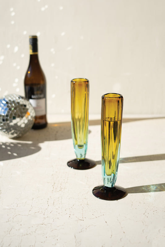 BlueandAmberRecycledChampagneGlass - Two yellow-tinted glasses on a reflective surface with a bottle and disco ball in the background.