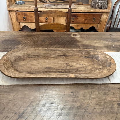An oval rustic wooden serving platter with a distressed finish, displayed on a matching rustic wooden table with a white runner. A wooden chair and antique cabinet are visible in the background.