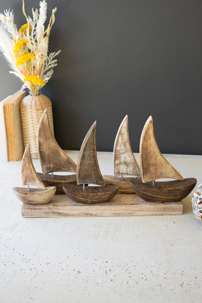 FiveWoodenSailboatsonaBase-1 - Wooden sailboat figurines on a stand with a vase of flowers in the background.