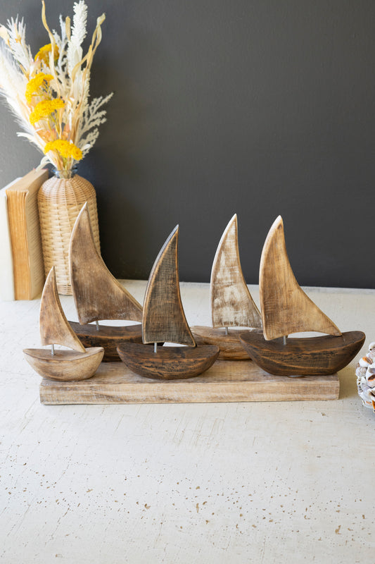 FiveWoodenSailboatsonaBase-1 - Wooden sailboat figurines on a stand with a vase of flowers in the background.