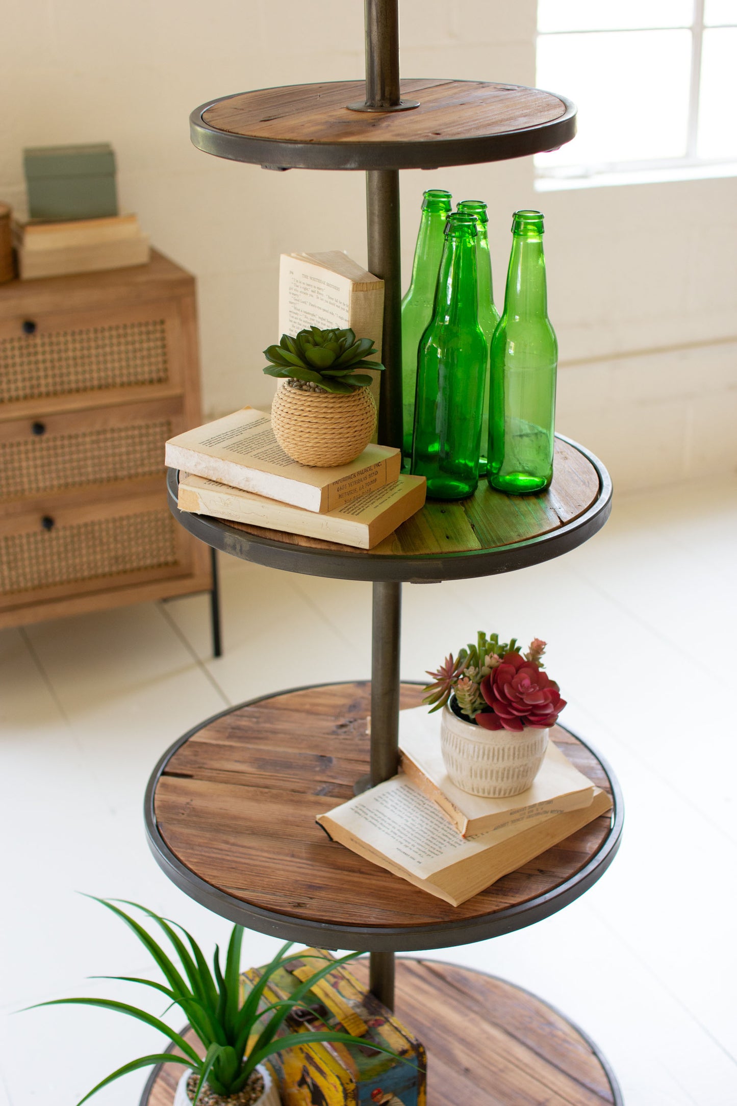 Four-tiered wooden and metal display shelf with books, plants, and green bottles.