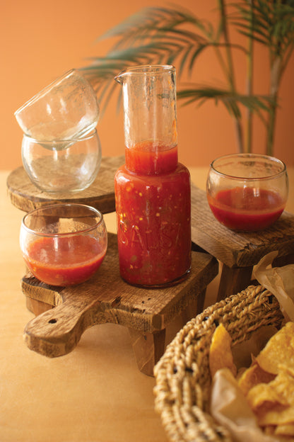 GlassSalsaPitcherwithFourBowls Glass containers with red sauce on a wooden stand with a plant in the background