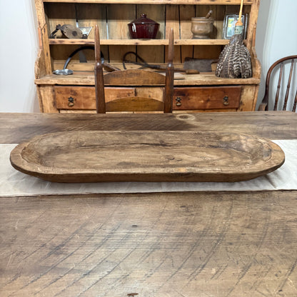 A rustic oval wooden serving tray with a natural wood grain finish, displayed on a matching wooden table in front of a wooden hutch.