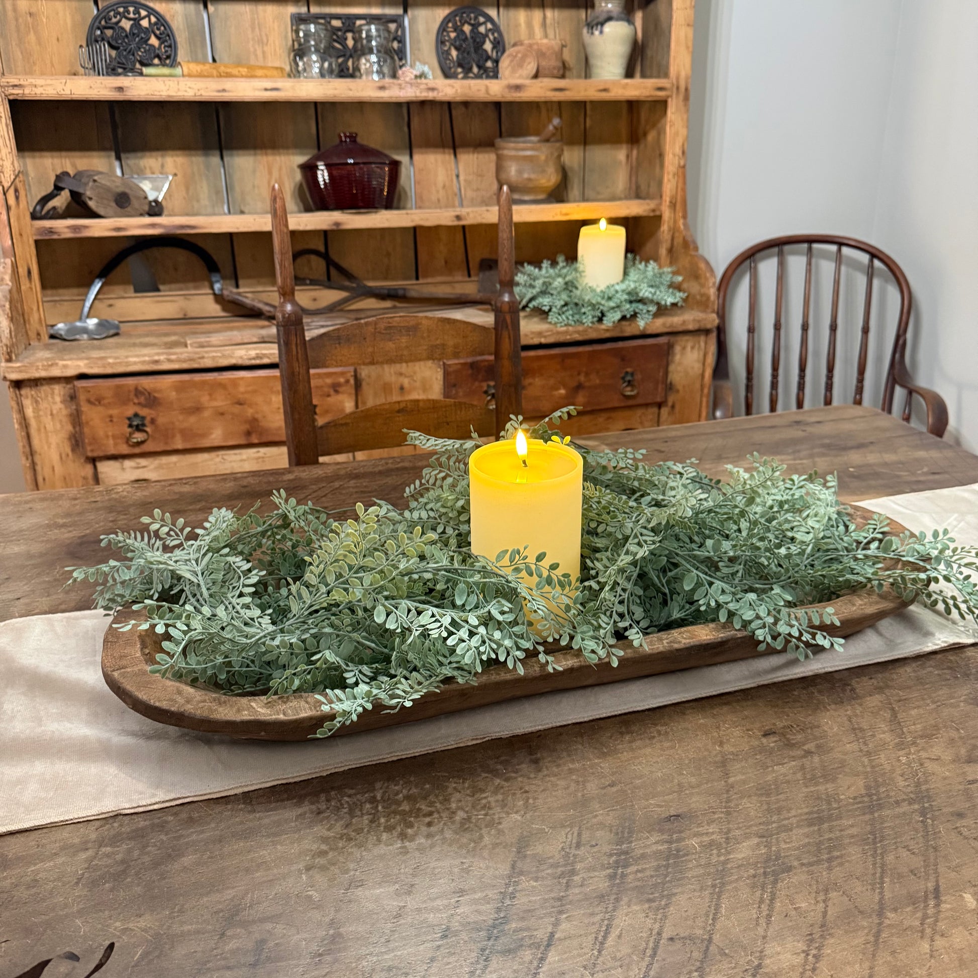 A rustic wooden tray centerpiece filled with green foliage and a yellow pillar candle, displayed on a wooden dining table in a home setting with a wooden hutch and chairs in the background.