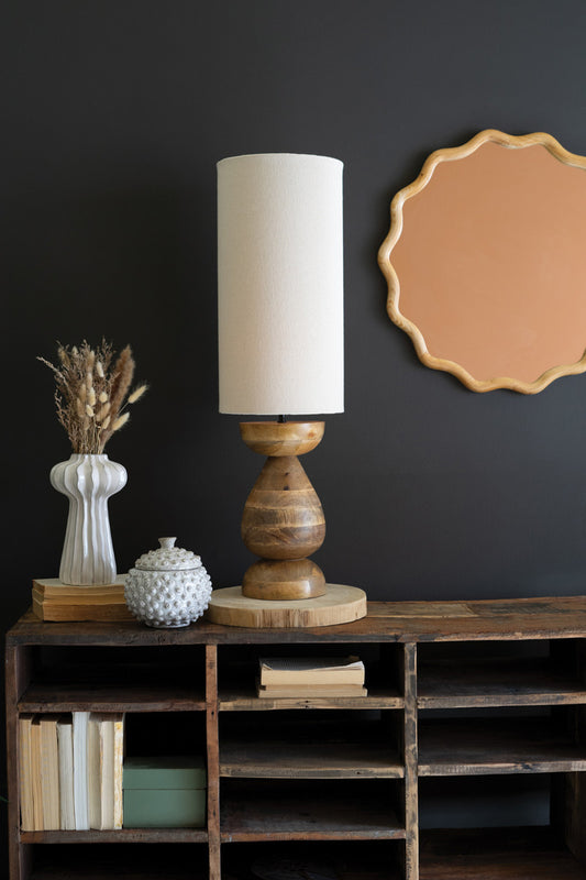 A mango wood table lamp base with a fabric cylinder lampshade, displayed on a wooden table with a dark background. The base is beige and the fabric shade is white.