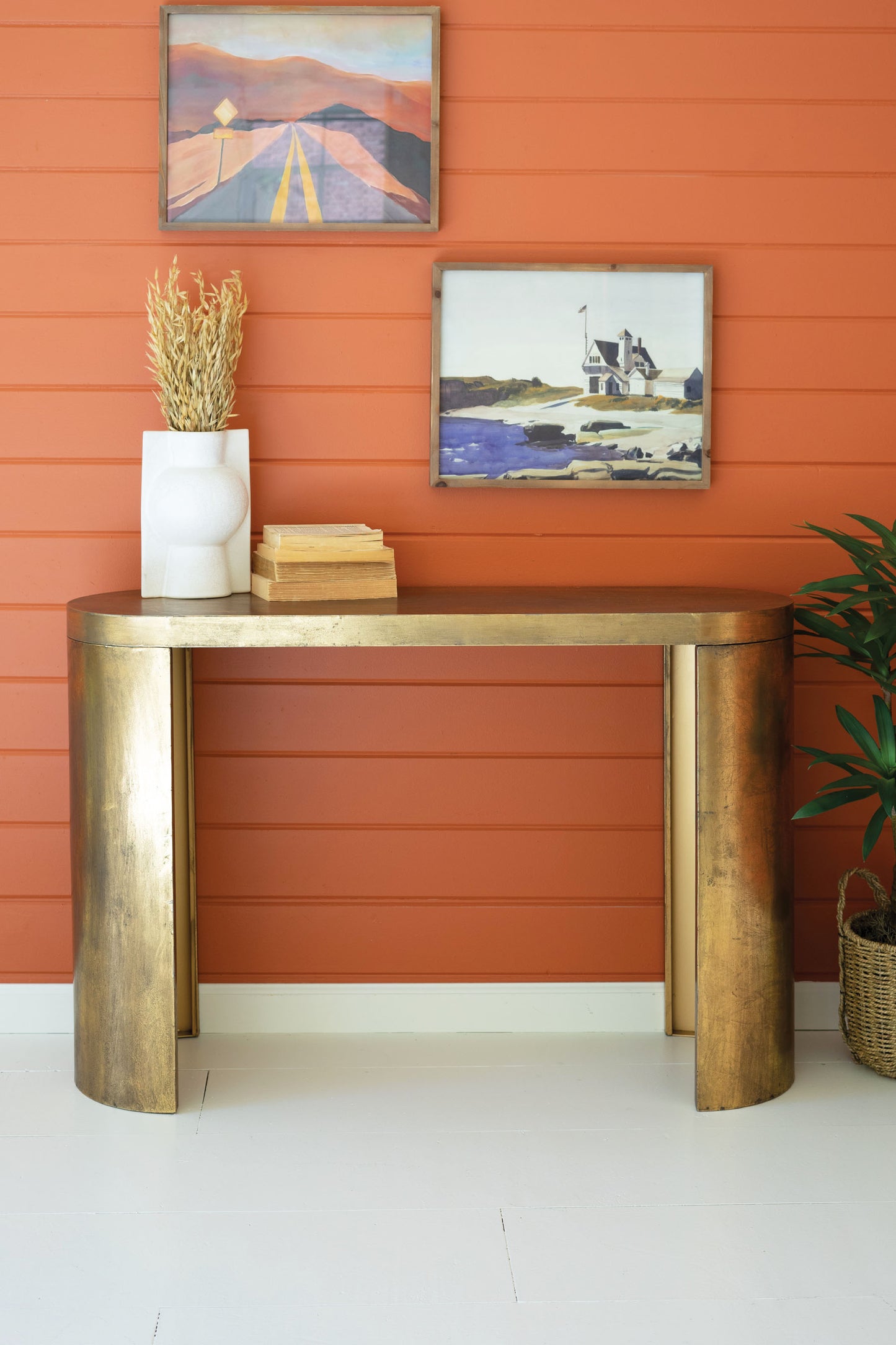 Oval Antique Brass Console Table against an orange wall with two framed artworks.