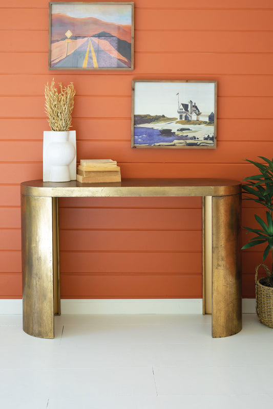 Oval Antique Brass Console Table against an orange wall with two framed artworks.