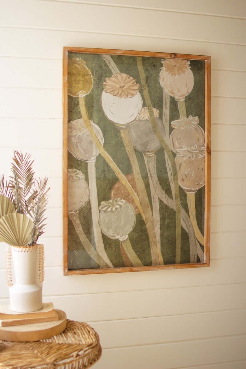A framed poppy print artwork in shades of green and brown, displayed on a wall above a wicker table.