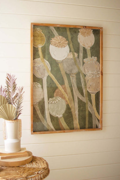 A framed poppy print artwork in shades of green and brown, displayed on a wall above a wicker table.