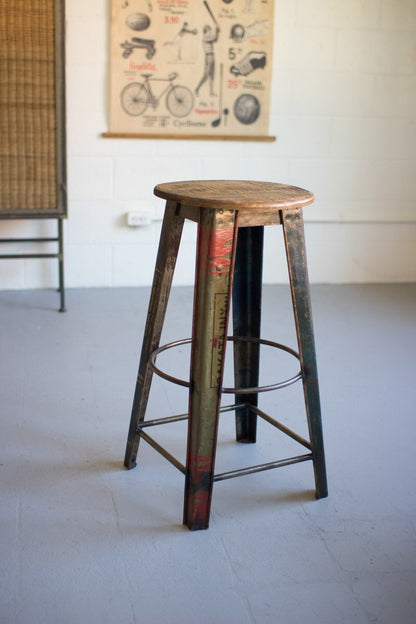 Vintage-style RecycledMetalBarStoolwithWoodenSeat in a room with a poster on the wall.