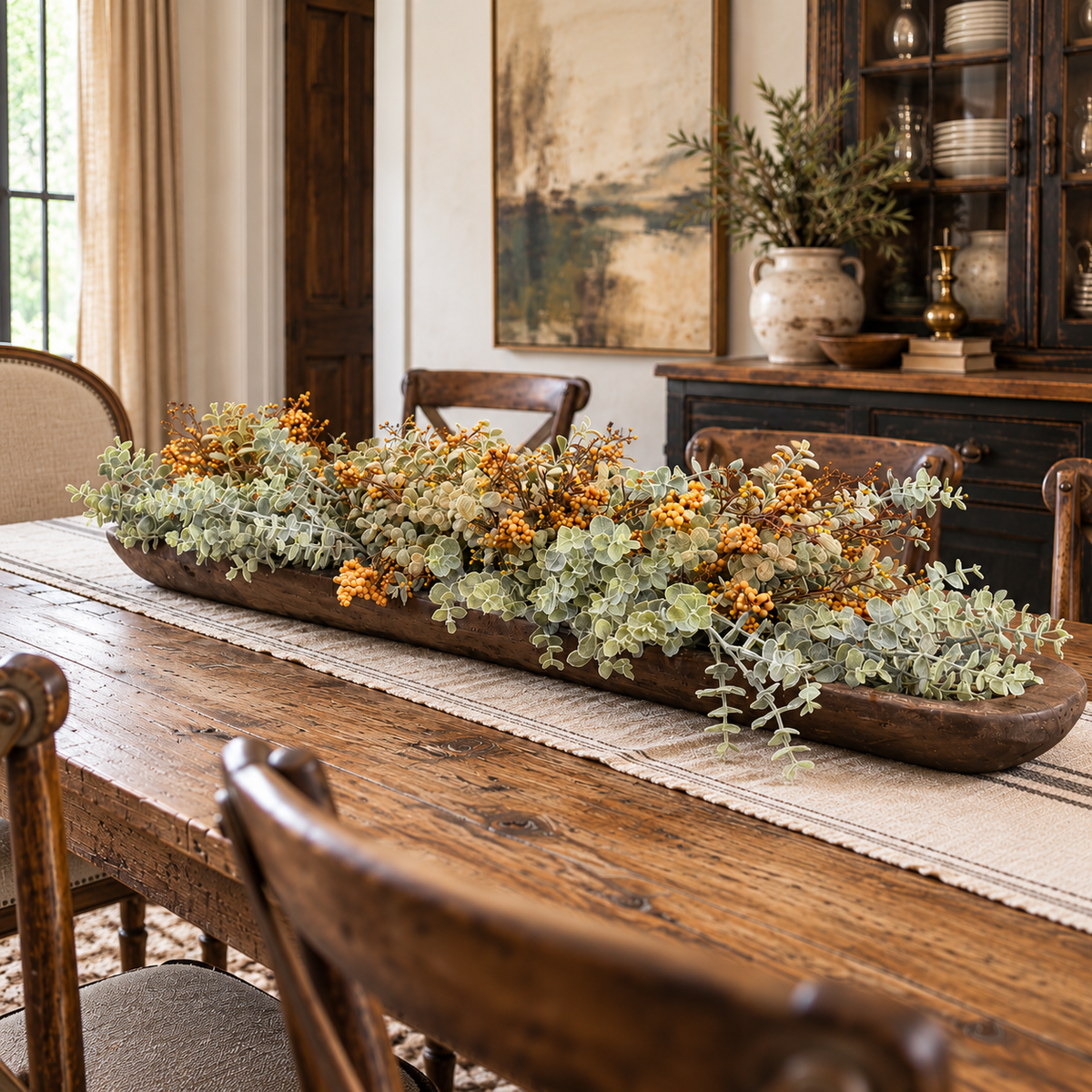 A long rustic wooden tray filled with dried orange berries, sage green eucalyptus, and other autumn foliage, styled on a dining table in a warm, traditional dining room.