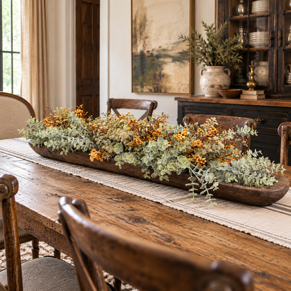 A long rustic wooden tray filled with dried orange berries, sage green eucalyptus, and other autumn foliage, styled on a dining table in a warm, traditional dining room.