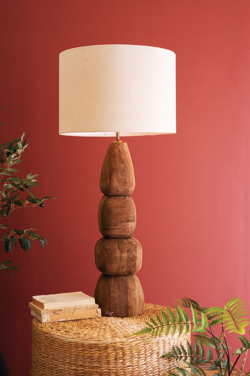 Rustic Mango Wood Table Lamp  a white shade on a woven surface against a red wall.