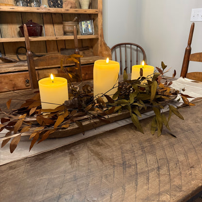 Three lit pillar candles arranged in a rustic wooden tray with dried leaves and greenery, set on a dining table in a home interior.