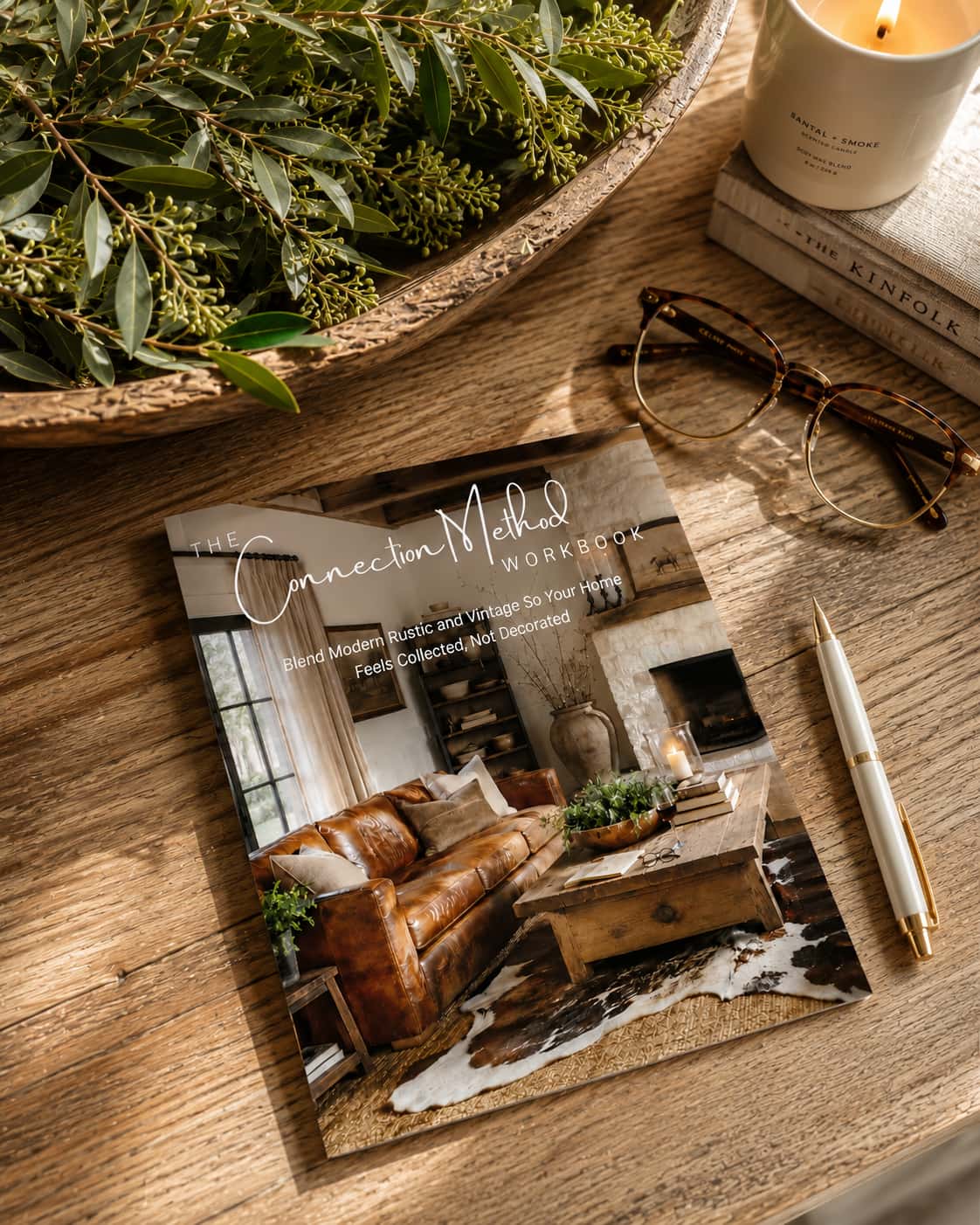 A printed card or booklet titled 'Connection Method WORKROOM' lies on a wooden table, styled with a candle, eyeglasses, a pen, stacked books, and greenery. The cover shows a warm living-room interior photo.