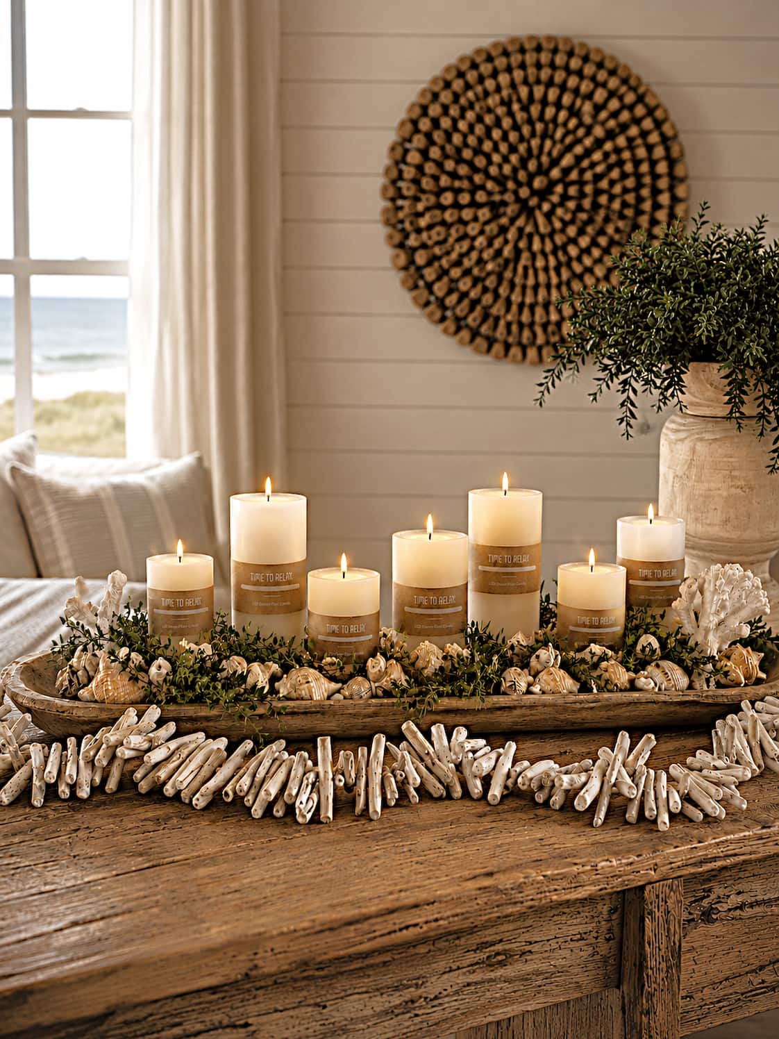 coastal style centerpiece with  candles, drifwood garland, and seashells and plants on a wooden surface in a room with a window and wall art.