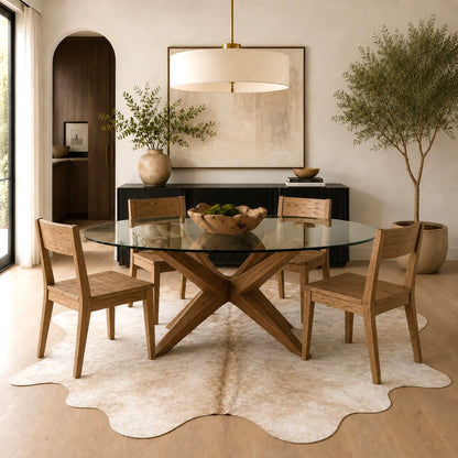 A modern dining room scene featuring a round glass-top table with a wooden X-shaped base, surrounded by four light wood chairs on a white cowhide rug. The room is styled with a black sideboard, a large canvas print, a pendant light, and potted trees.
