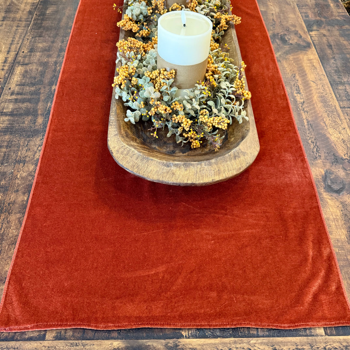 A rust-red velvet table runner laid out on a dark wood table, styled with a wooden tray, a white pillar candle, and a dried floral arrangement.
