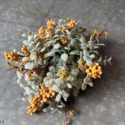 A cluster of artificial decorative foliage with dusty sage-green leaves and small round golden-orange berries, arranged on a gray stone-like surface.