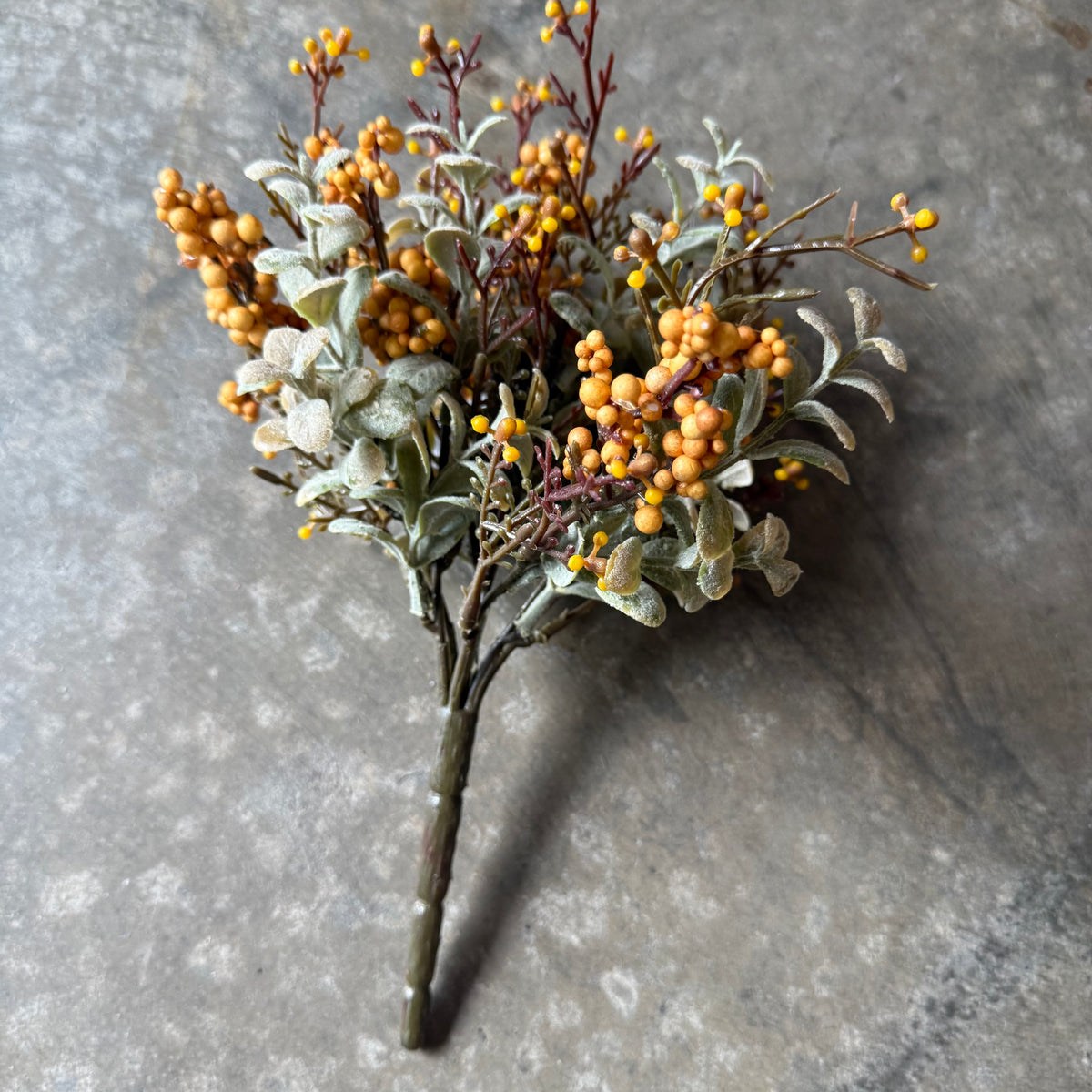 An artificial floral arrangement featuring clusters of small orange berries, dusty silver-gray leaves, and dark brown stems, displayed on a textured gray stone surface.