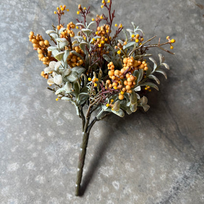 An artificial floral arrangement featuring clusters of small orange berries, dusty silver-gray leaves, and dark brown stems, displayed on a textured gray stone surface.