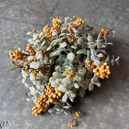 A cluster of artificial floral stems featuring dusty sage-green leaves and small round golden-yellow berries, arranged on a textured gray stone surface.