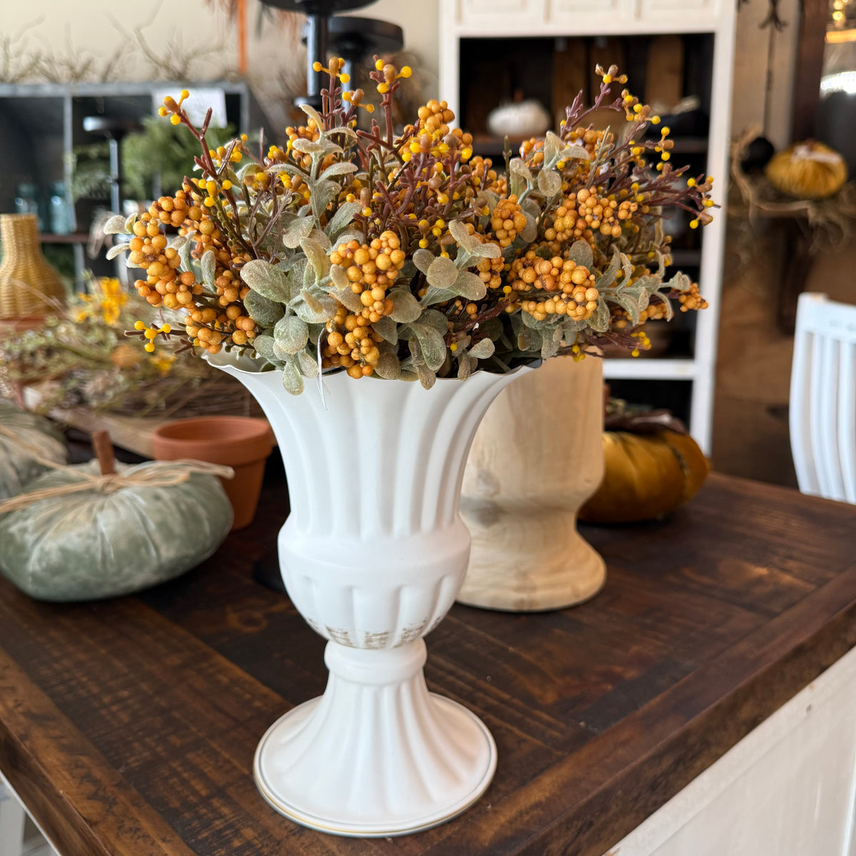 A white fluted pedestal vase filled with artificial autumn sprigs featuring small orange berries and muted green-gray leaves, displayed on a dark wood table in a home decor store.