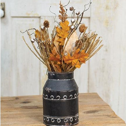 A rustic black metal milk can vase filled with dried autumn foliage, including orange leaves, brown pinecones, and tan grasses, displayed on a wooden table against a white paneled wall.