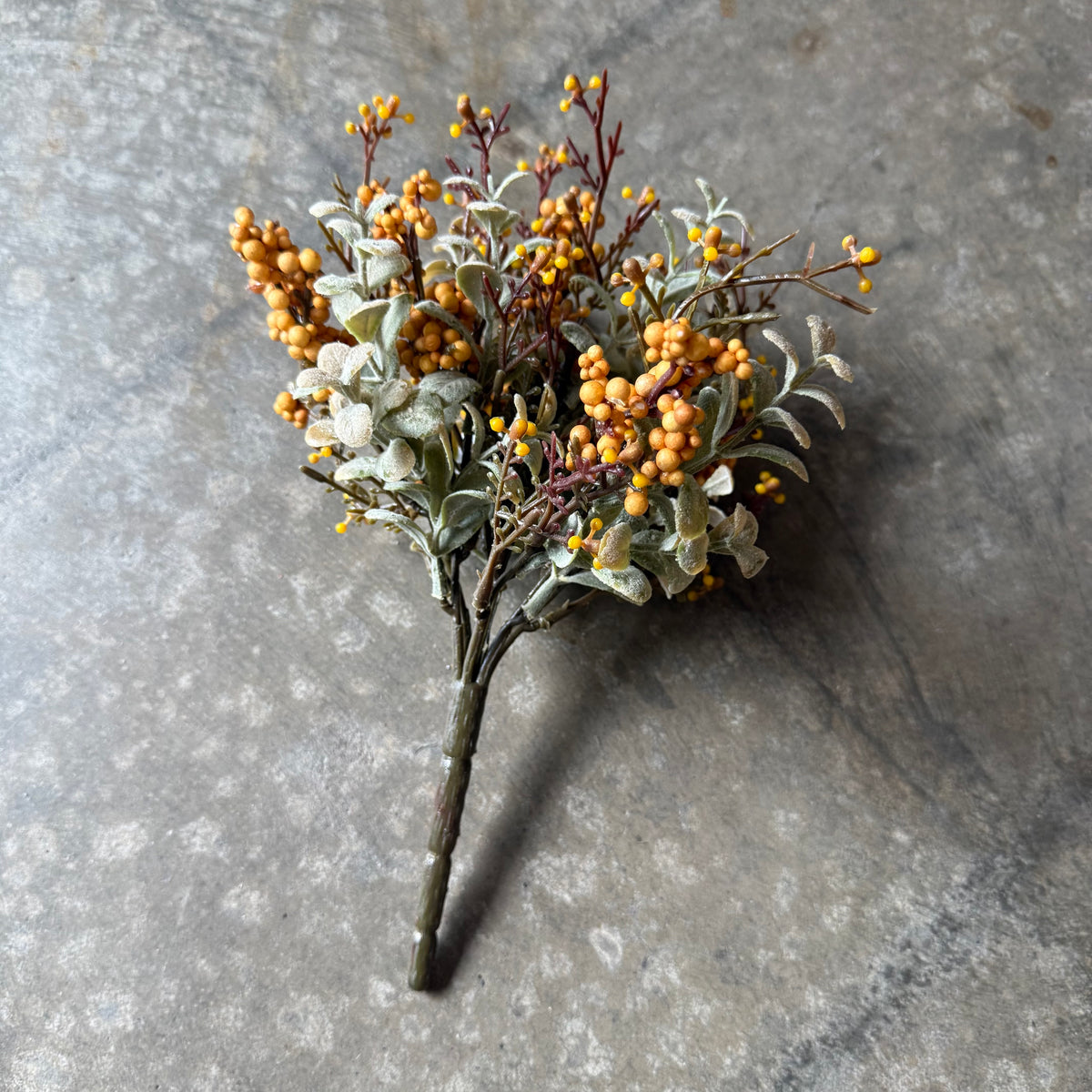 An artificial floral arrangement of yellow berries, dusty white leaves, and brown stems, photographed on a textured gray stone surface.