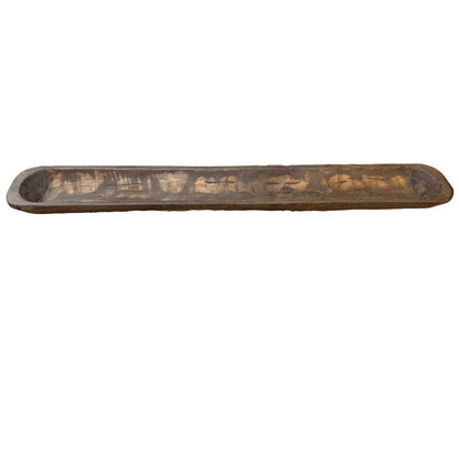 A long, narrow rustic wooden bowl with a dark brown finish and visible grain, displayed on a patterned runner.