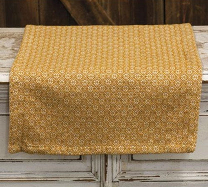 A rectangular table runner with a textured mustard yellow and white geometric pattern, draped over a rustic white wooden table.