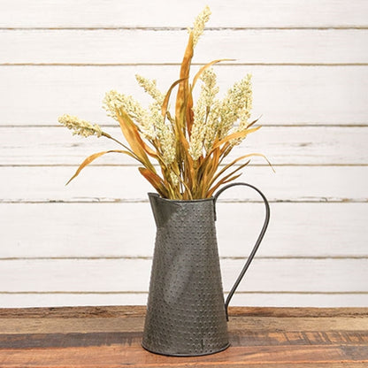 A rustic metal pitcher vase filled with dried wheat and grass stems, set on a wooden surface against a white wood plank wall.