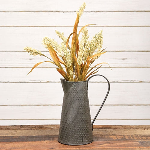 A rustic metal pitcher vase filled with dried wheat and grass stems, set on a wooden surface against a white wood plank wall.