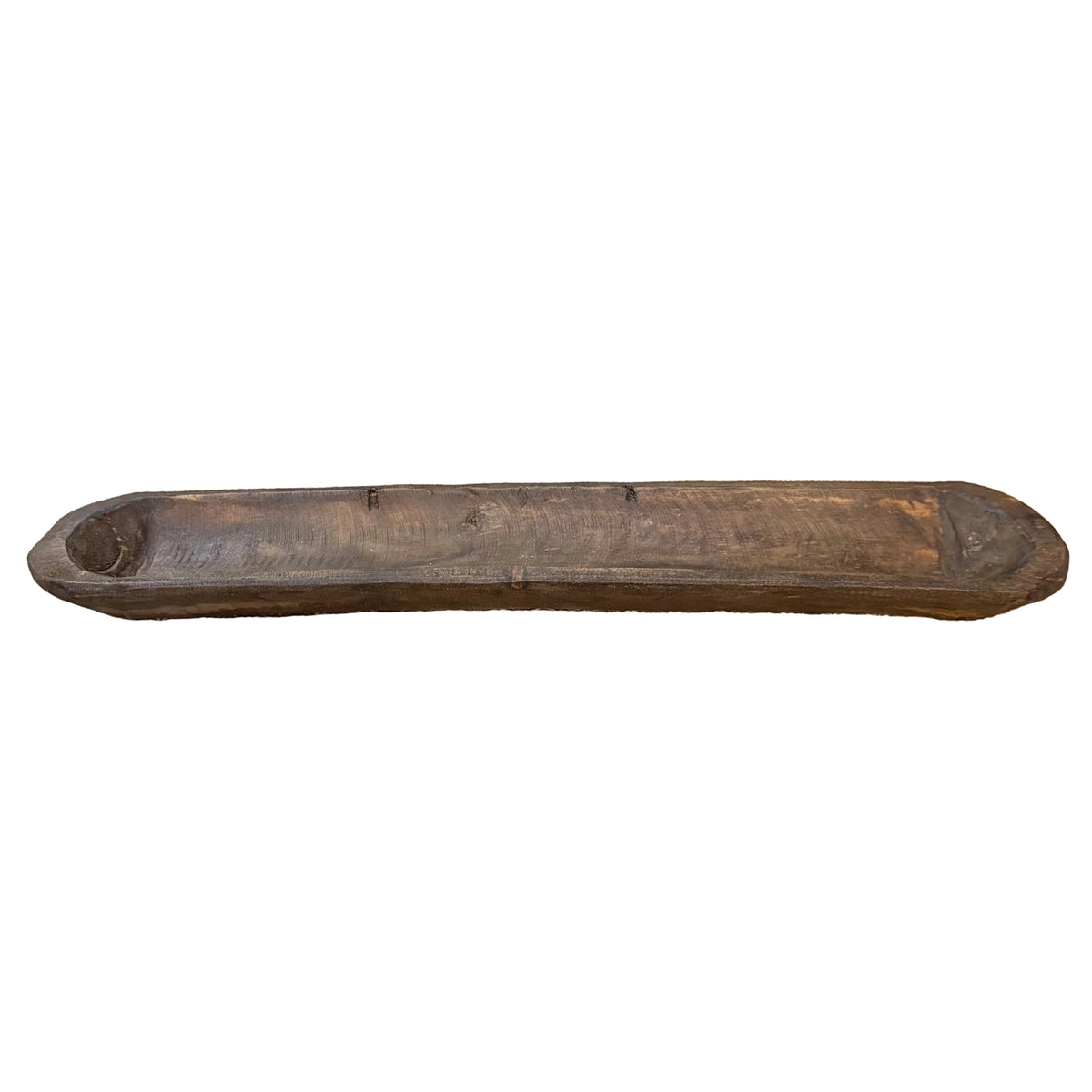 A long, rustic carved wooden bowl, displayed on a patterned runner on a wooden table with a wooden cabinet in the background.