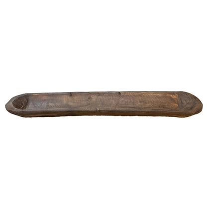 A long, rustic carved wooden bowl, displayed on a patterned runner on a wooden table with a wooden cabinet in the background.