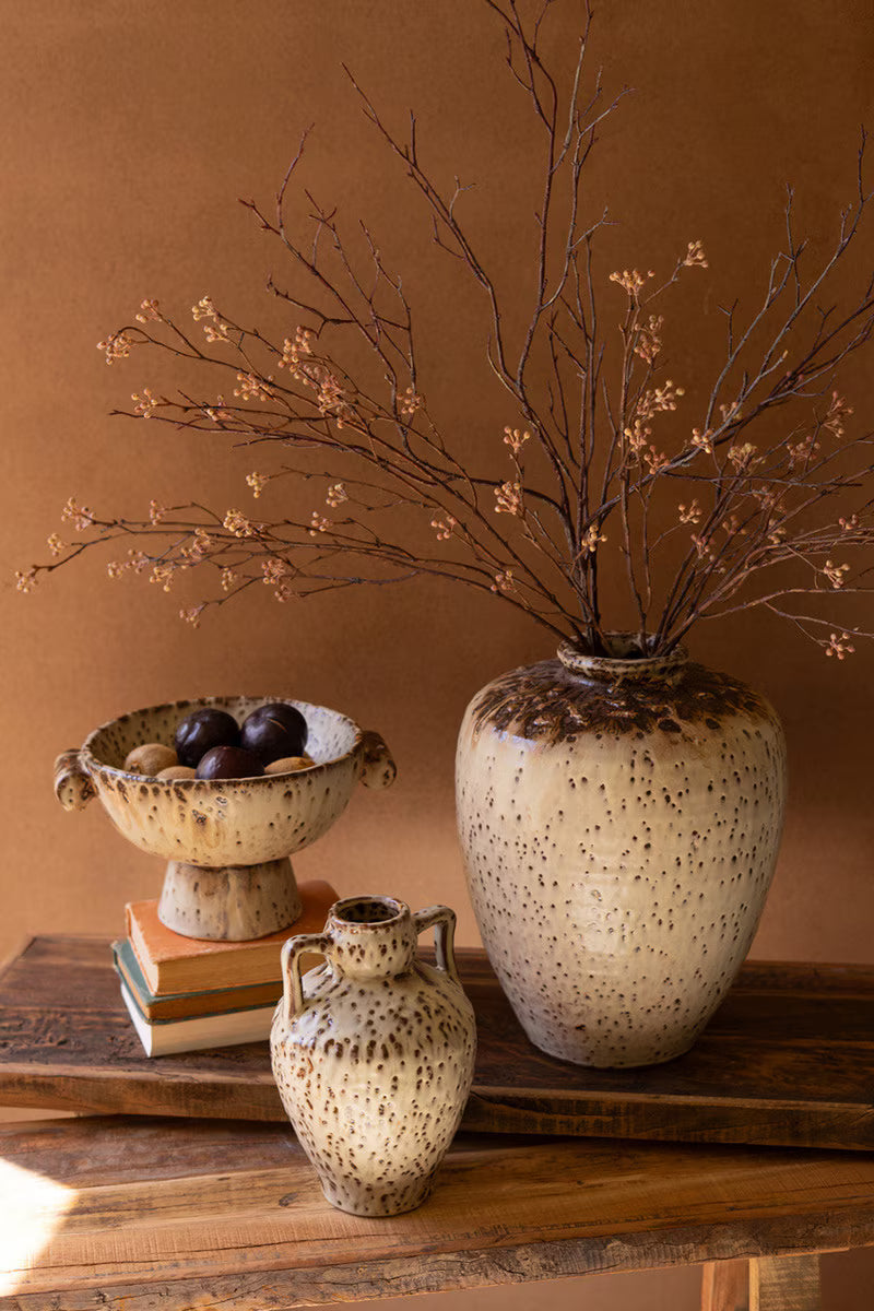 Decorative setting with speckled vases and a branch arrangement on a wooden surface against a brown background.