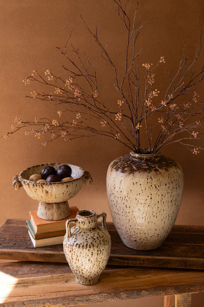 Decorative setting with speckled vases and a branch arrangement on a wooden surface against a brown background.