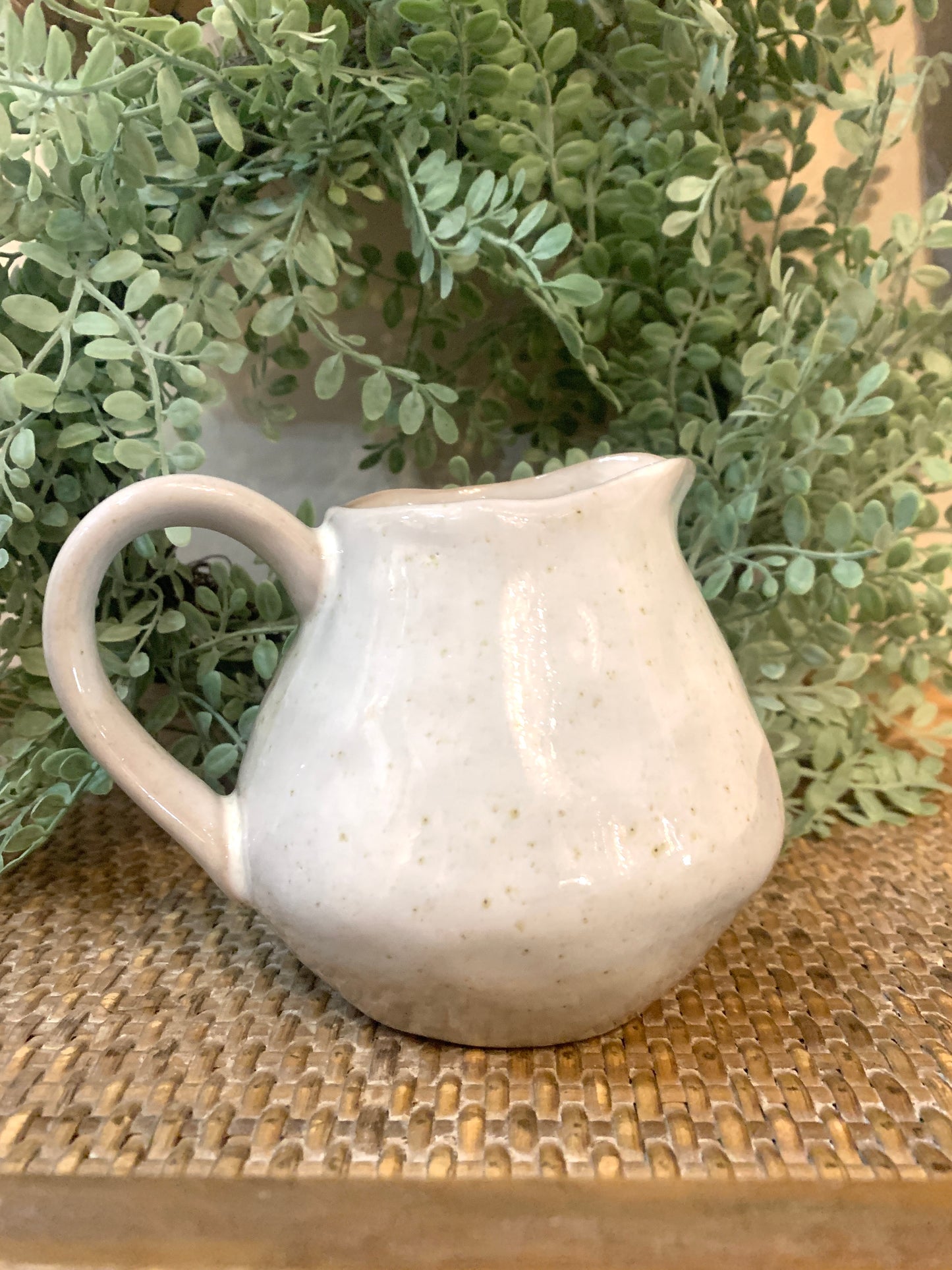 A white ceramic pitcher placed on a woven surface with a green plant in the background.