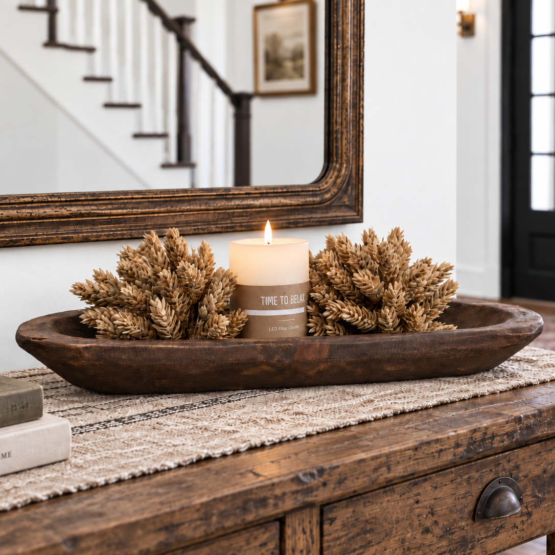A rustic dark brown wooden tray holding two dried wheat-like decorative pieces and a lit cream candle on  a wooden console table in a home entryway.