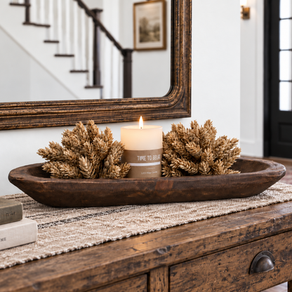 A rustic dark brown wooden tray holding two dried wheat-like decorative pieces and a lit cream candle on  a wooden console table in a home entryway.